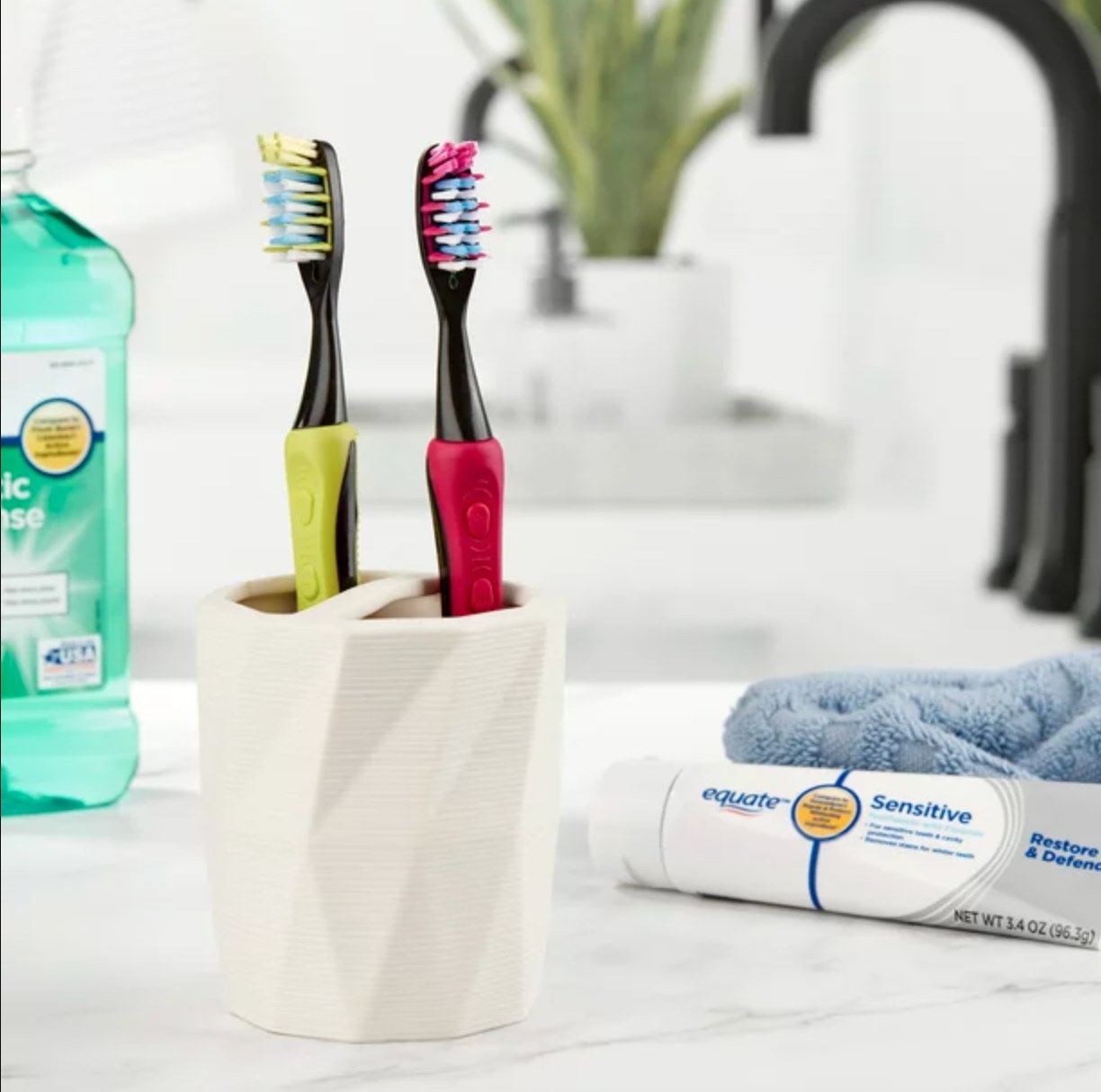 the green and pink electric toothbrushes in a cup on a counter with toothpaste and mouthwash