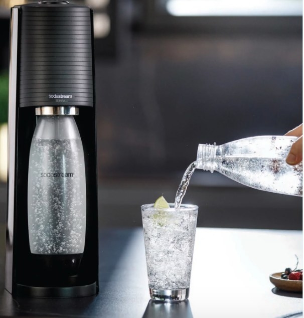 a model pouring sparkling water into a glass next to a sodastream machine with another carafe of sparkling water in it