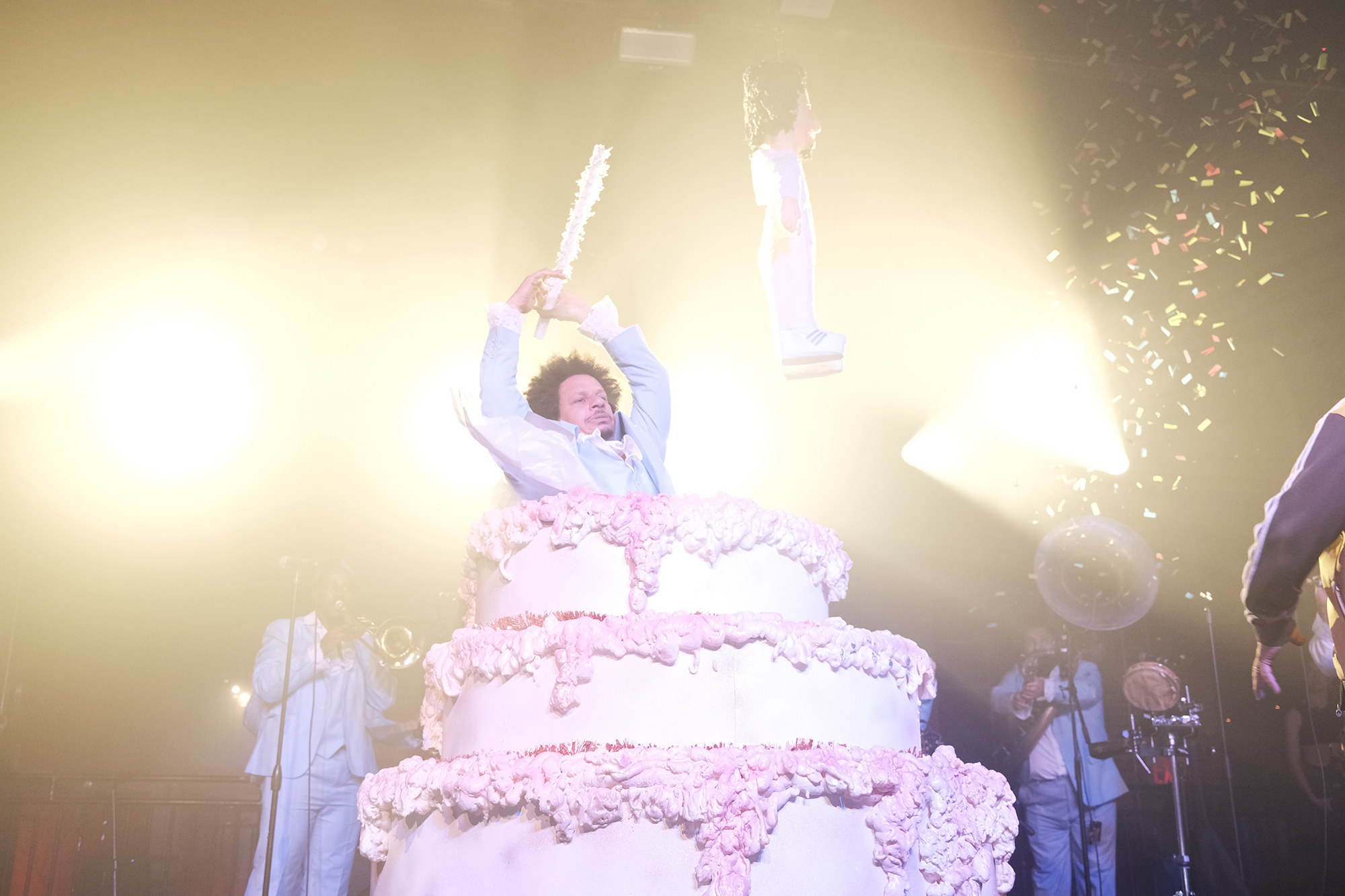 Eric André emerging with his arms raised from a gigantic pink cake