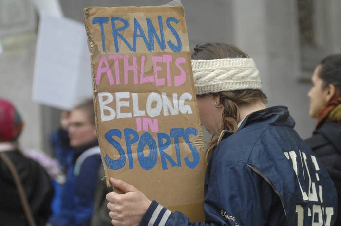 A protester holds up a cardboard sign reading &quot;Trans athletes belong in sports&quot; spelled out in blue, pink, and white, the colors of the trans flag