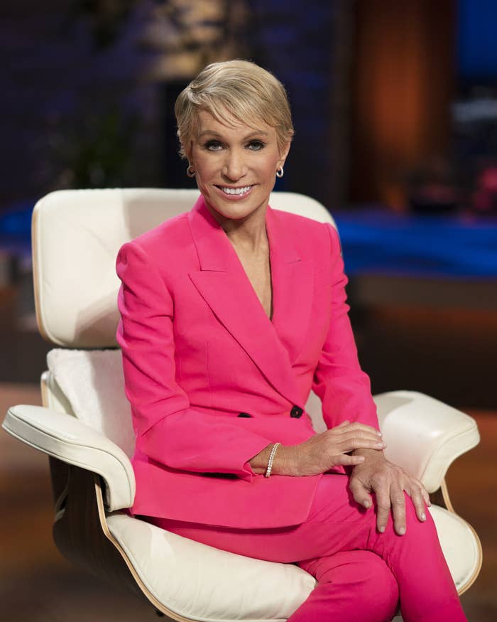 Barbara smiles at the camera as she sits on the set of Shark Tank. She&#x27;s wearing a tailored pantsuit and simple jewelry