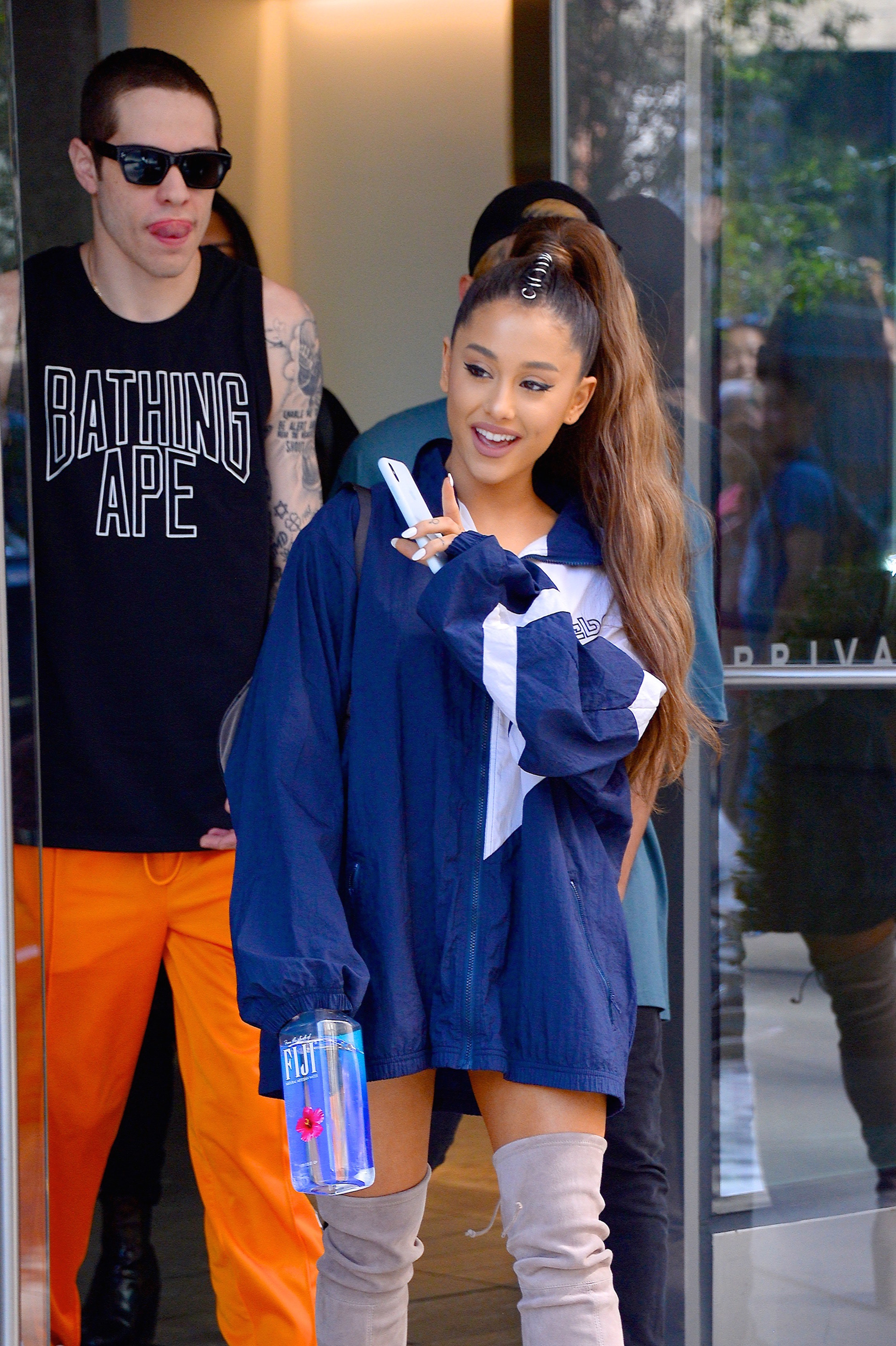 Ariana Grande, wearing an oversize men&#x27;s jacket, walks out of a building waving, Pete Davidson is walking behind her