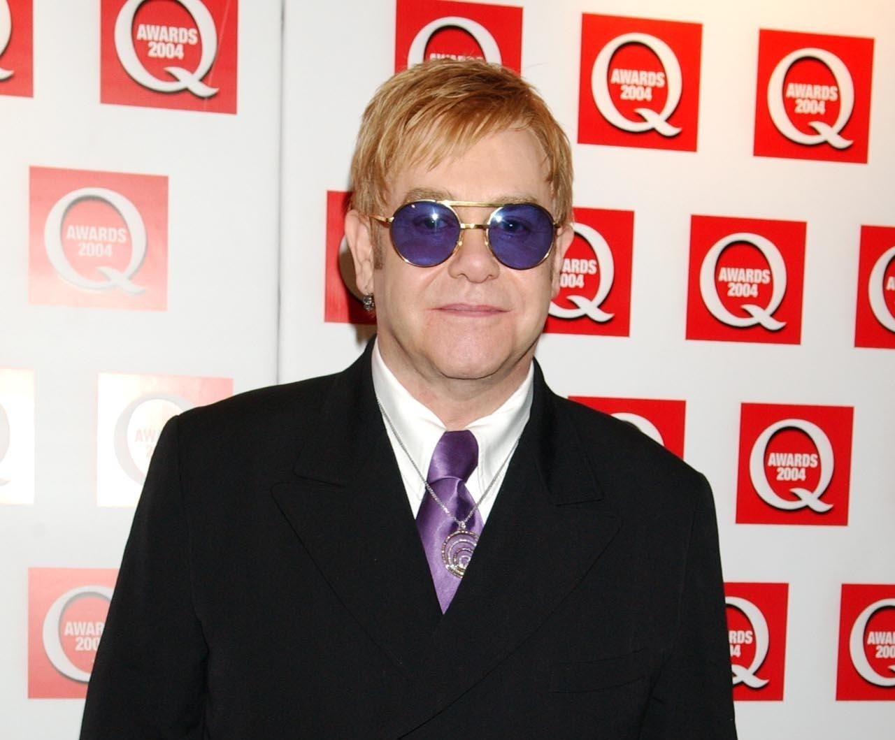 Elton John, in a black suit with purple tie and purple sunglasses, poses on the red carpet of the 2004 Q Awards