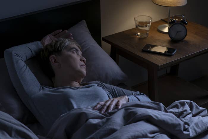 A woman lying awake in bed next to a bedside table that has an old-school alarm clock, a cellphone, and a glass of water