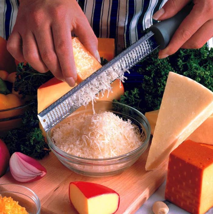 a model using the microplane to great cheese into a bowl on a counter surrounded by cheeses and vegetables