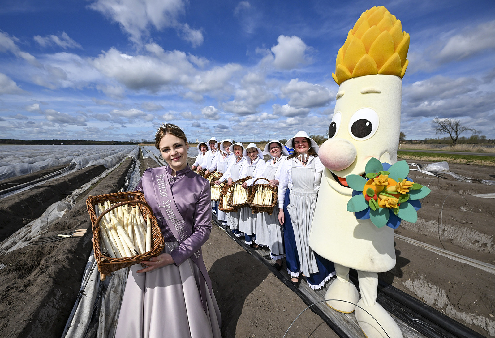 the asparagus queen stands holding a basket of asparagus next to an asparagus mascot and other women holding asparagus