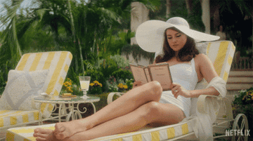 A white woman in an old fashioned bathing suit and large sunhat sits on a pool chair and reads a book