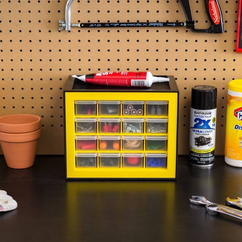 a yellow and black parts cabinet holding nails, batteries, and tool bits on a garage workbench next to tools and sprays