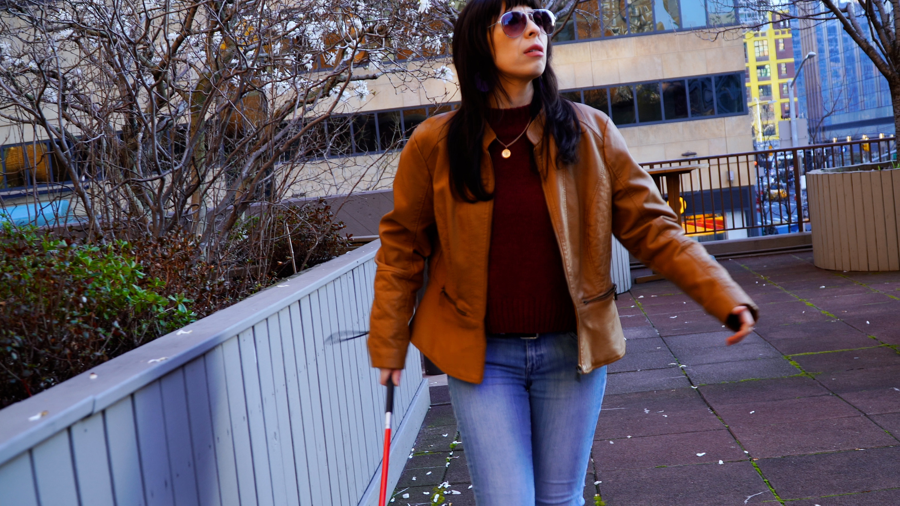 Logan (Melissa Jennifer Gonzalez) walks on an outdoor patio with a blind cane. They are wearing blue jeans, a maroon sweater, and a tan leather jacket. Logan is Latinx, female presenting.