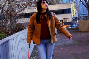 A Latinx female presenting person walks across an outdoor patio with a blind cane and sunglasses, pretending to be blind