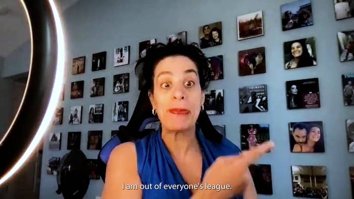 Maysoon Zayid as Yolanda, an olive-skinned woman with black hair wearing a blue blouse, points to a photo of younger her and Dave Matthews on a light blue wall filled with photos