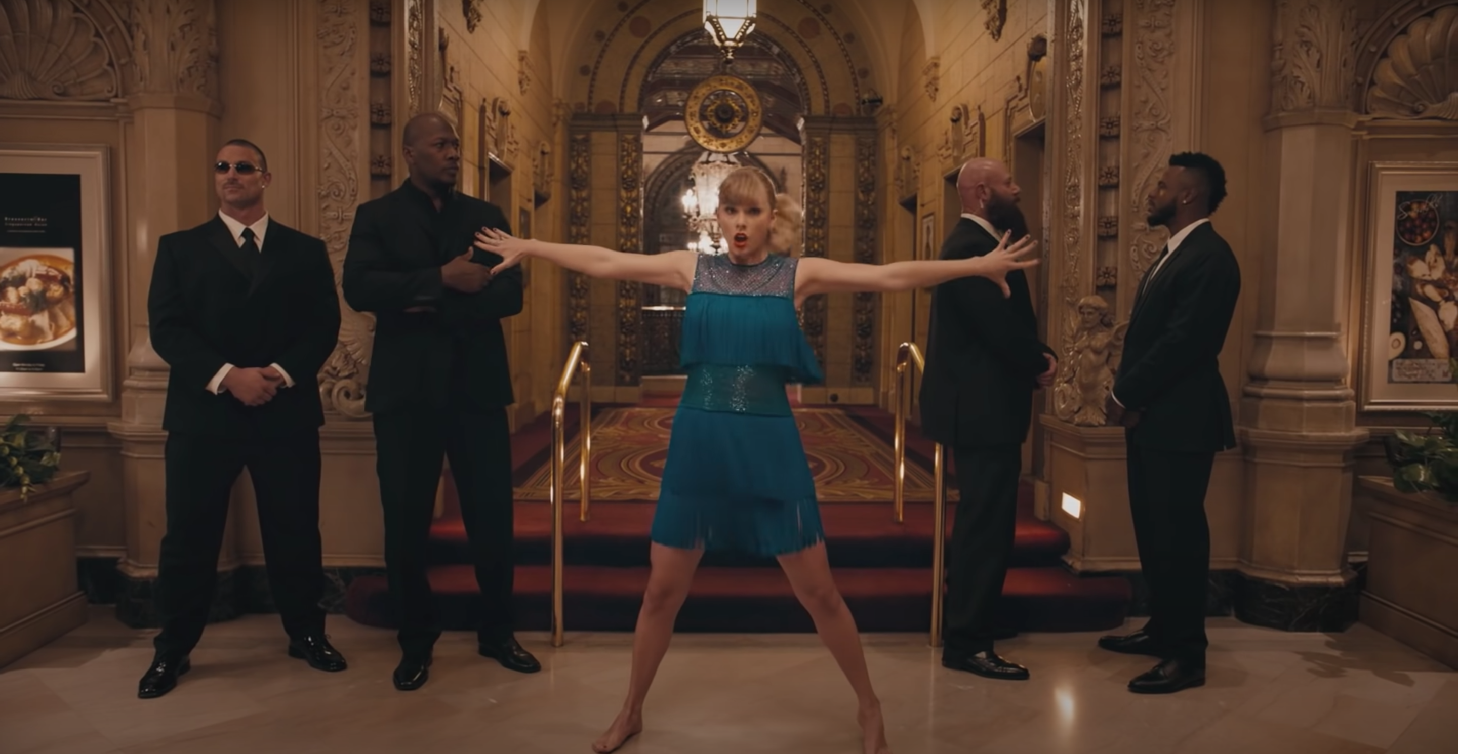 Taylor Swift wearing a blue dress in a hotel lobby with her arms outstretched and security guards on both sides.