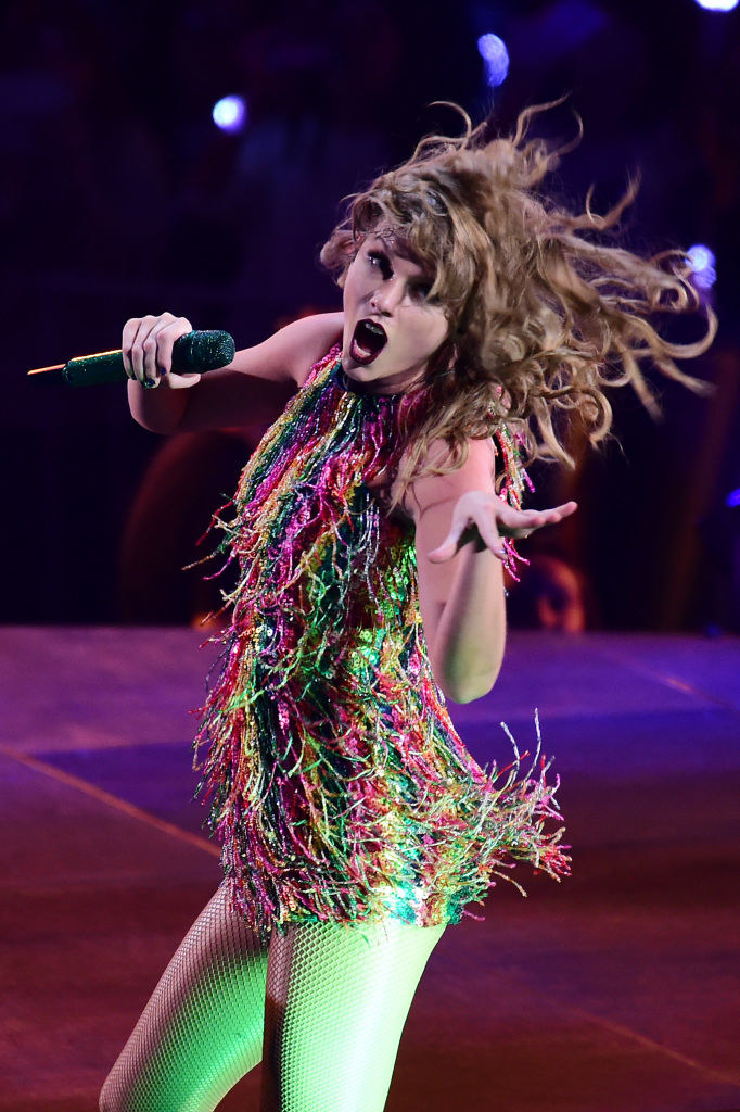 Taylor Swift wearing a colorful fringe dress with fishnet tights and singing into a microphone