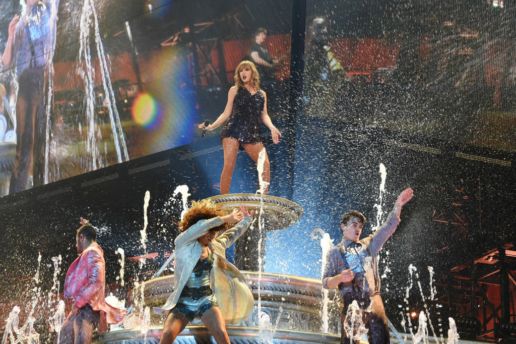 Taylor Swift on top of a fountain on a stage with dancers around her.