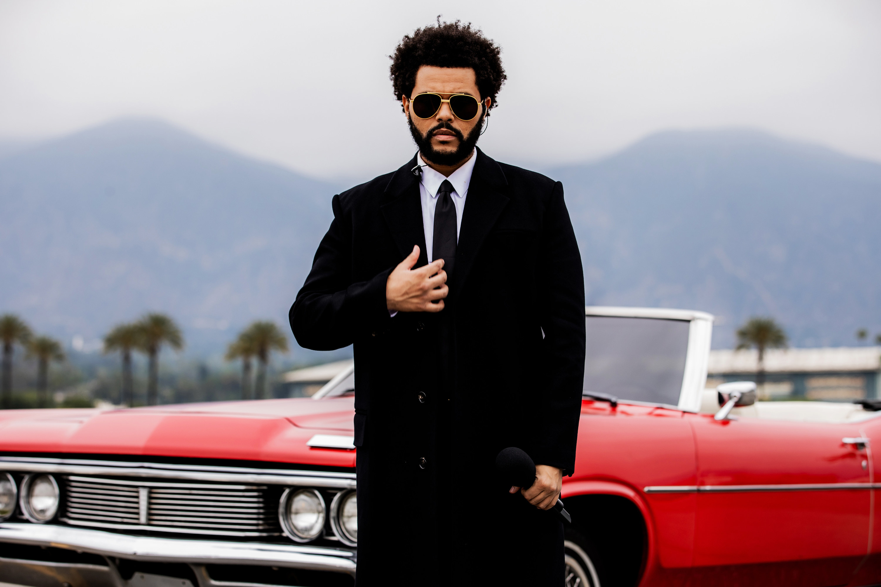 The Weeknd in a suit and coat and sunglasses with a convertible car behind him