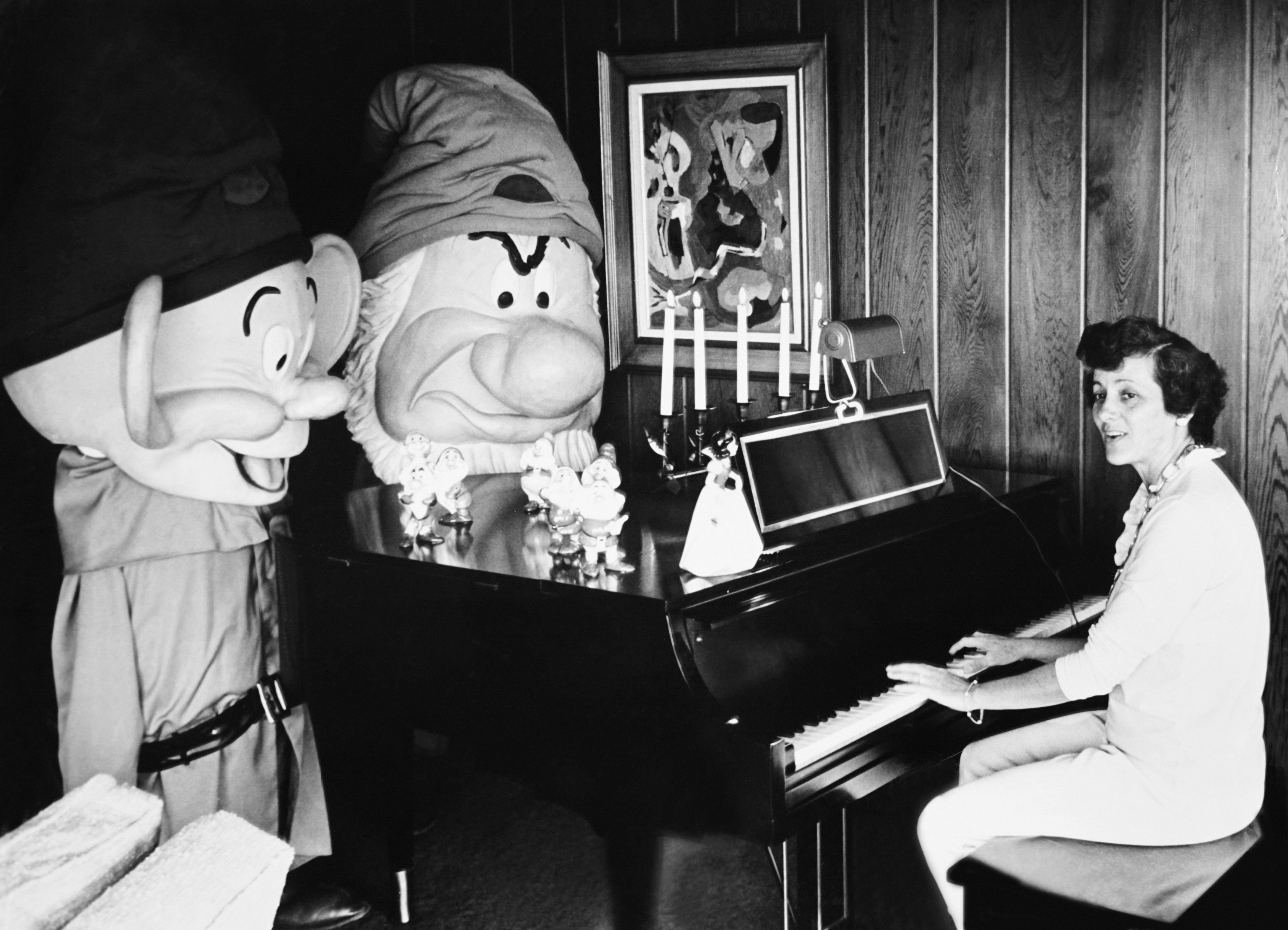 A woman playing piano with two people dressed as Snow White&#x27;s dwarfs standing next to the piano
