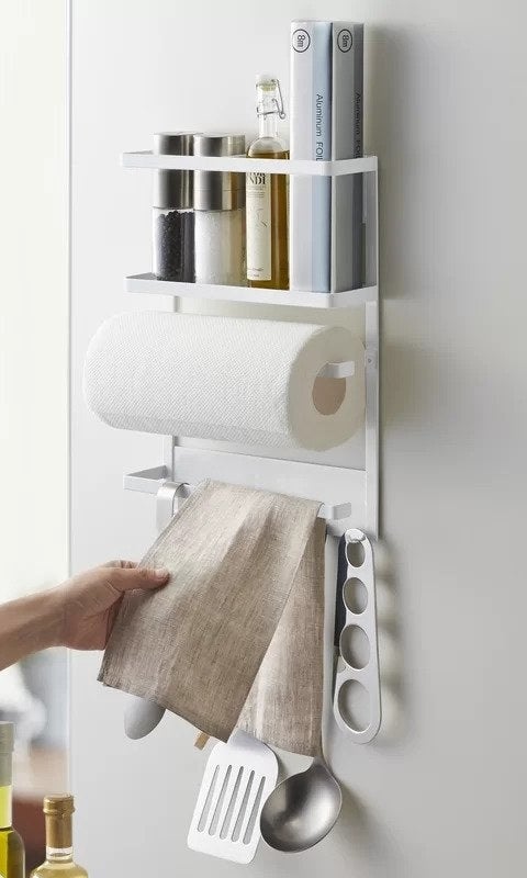 White magnetic rack with salt and pepper, bottle, and aluminum foil on top shelf, paper towels on middle rack, beige dish towel and utensils hanging from bottom rack
