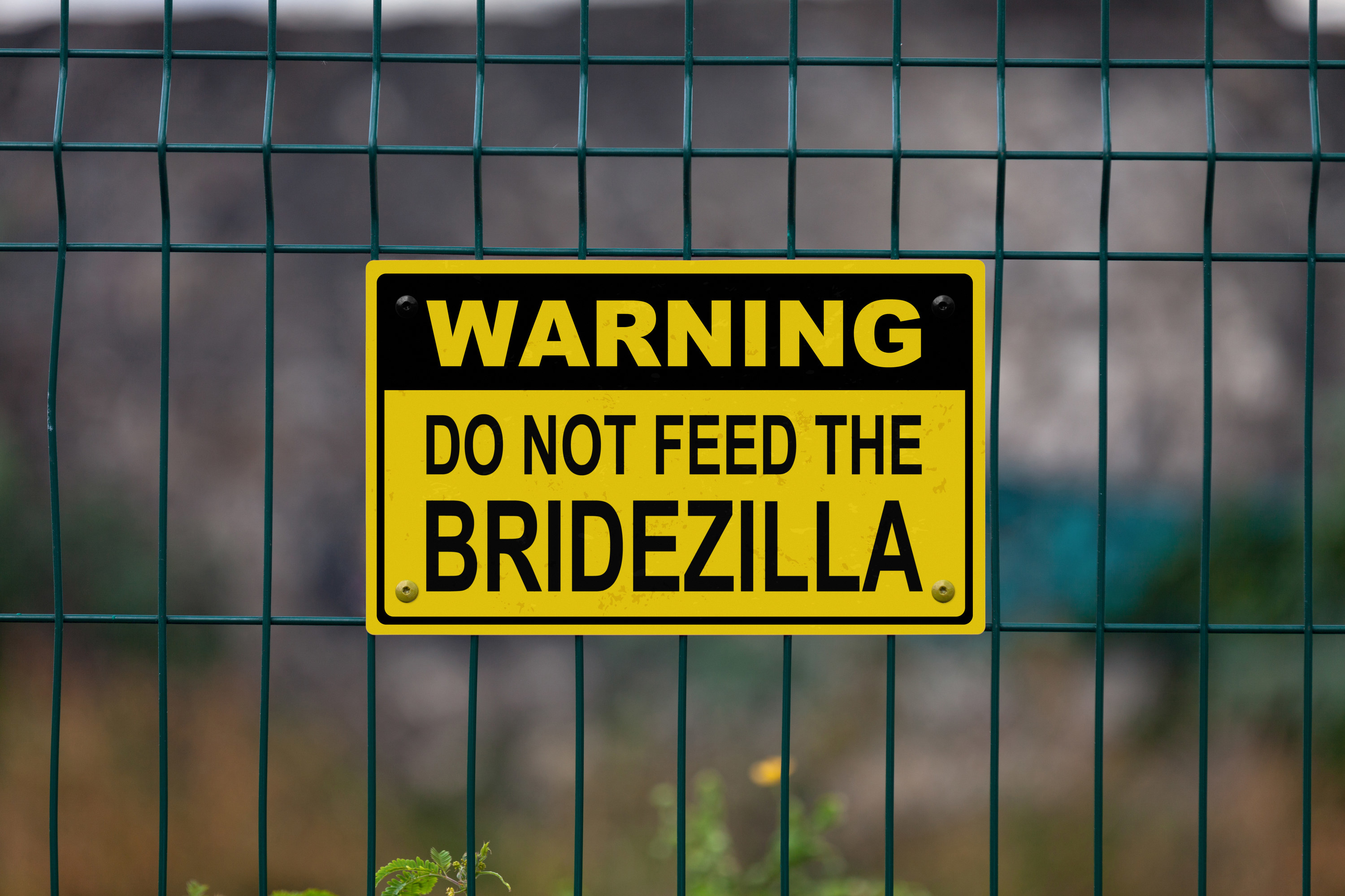 A yellow and black sign posted on a wire fence that reads &quot;Warning Do Not Feed The Bridezilla.&quot;