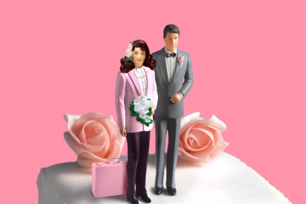 bride and groom figurines sitting on top of a wedding cake; the bride has a pink briefcase next to her