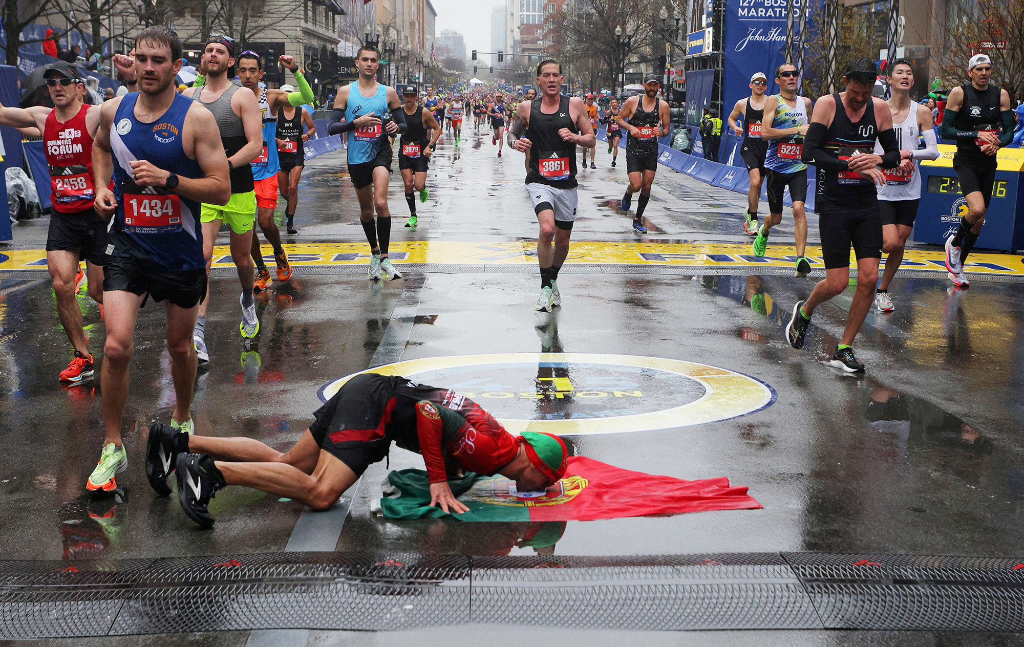 a man lies face down in a wet street  and kisses a Portuguese flag after running the boston marathon, behind him are many other runners