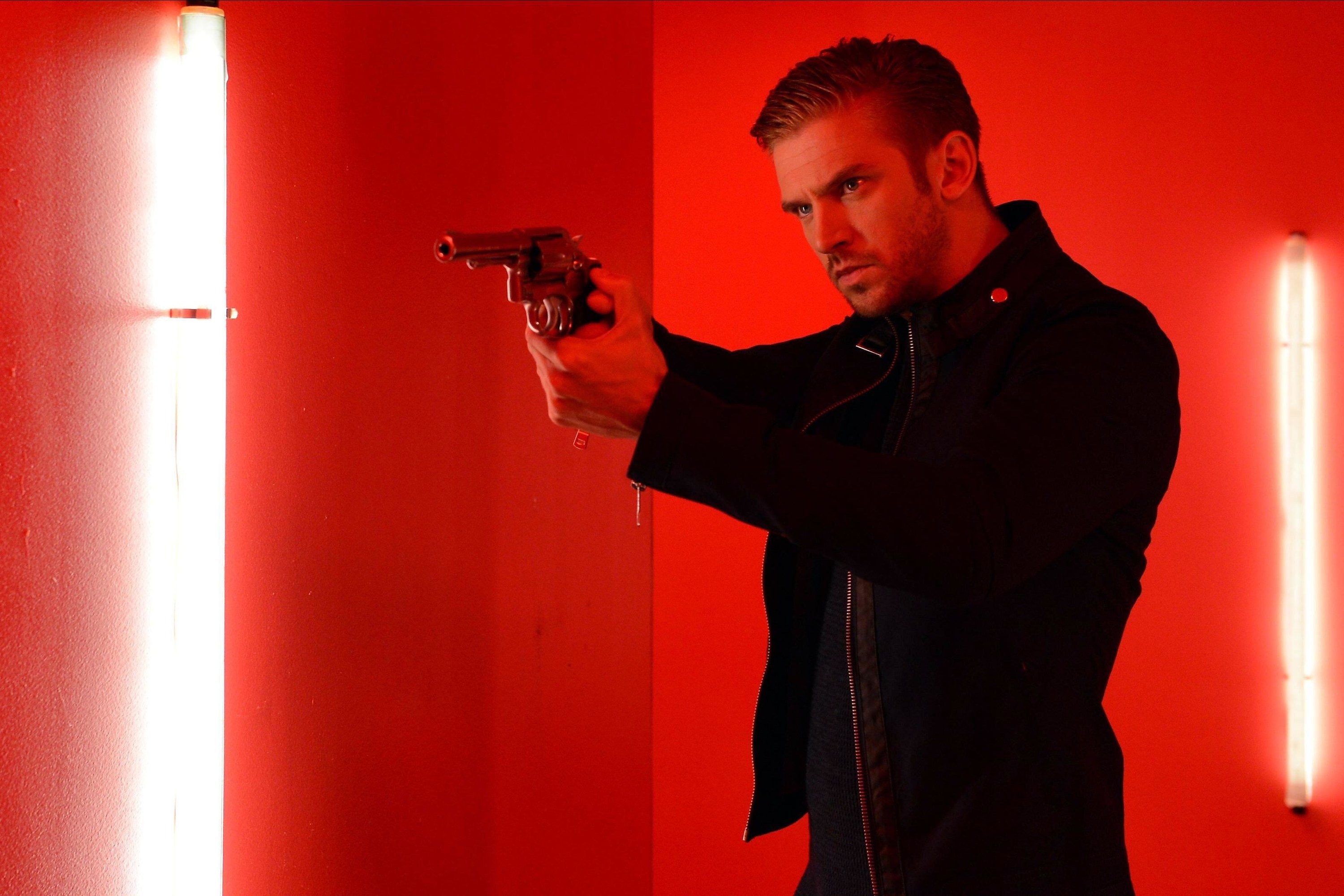 Dan Stevens holds a revolver in a bright red room