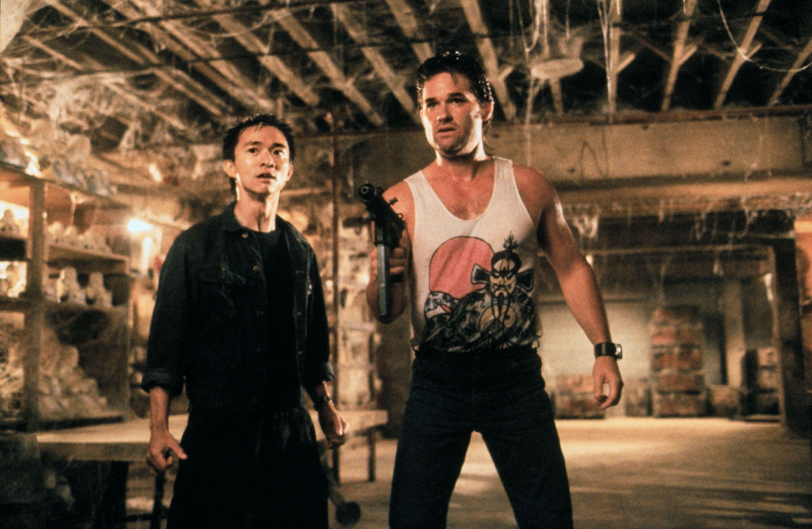 Dennis Dun and Kurt Russell stand with a gun in a dirty basement