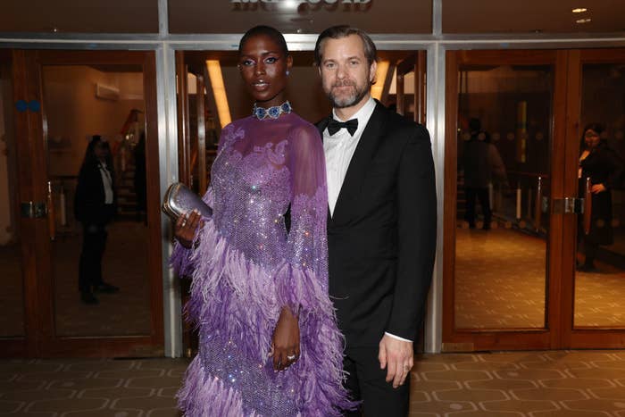 Jodie and Joshua pose together for an event photo in front of a building. Jodies is wearing a sheer and feathered gown and Joshua is wearing a suit and bowtie