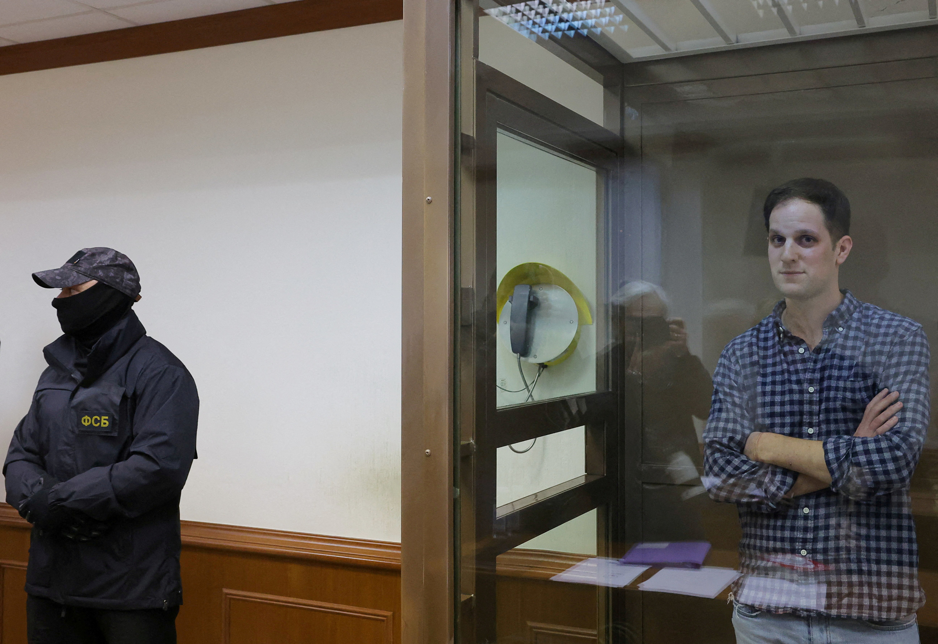 a man with crossed arms stands in a glass box next to a security guard