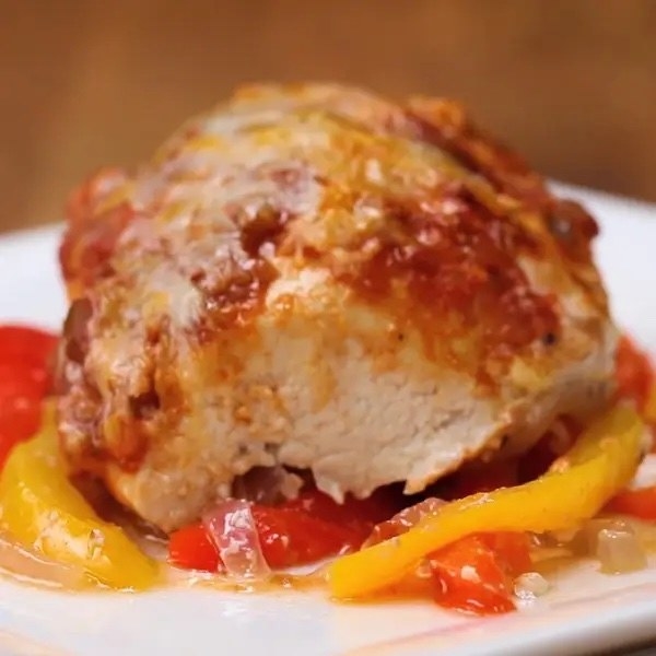 Closeup of a baked chicken breast topped with melted cheese and tomato sauce, served on a bed of sliced peppers and onions