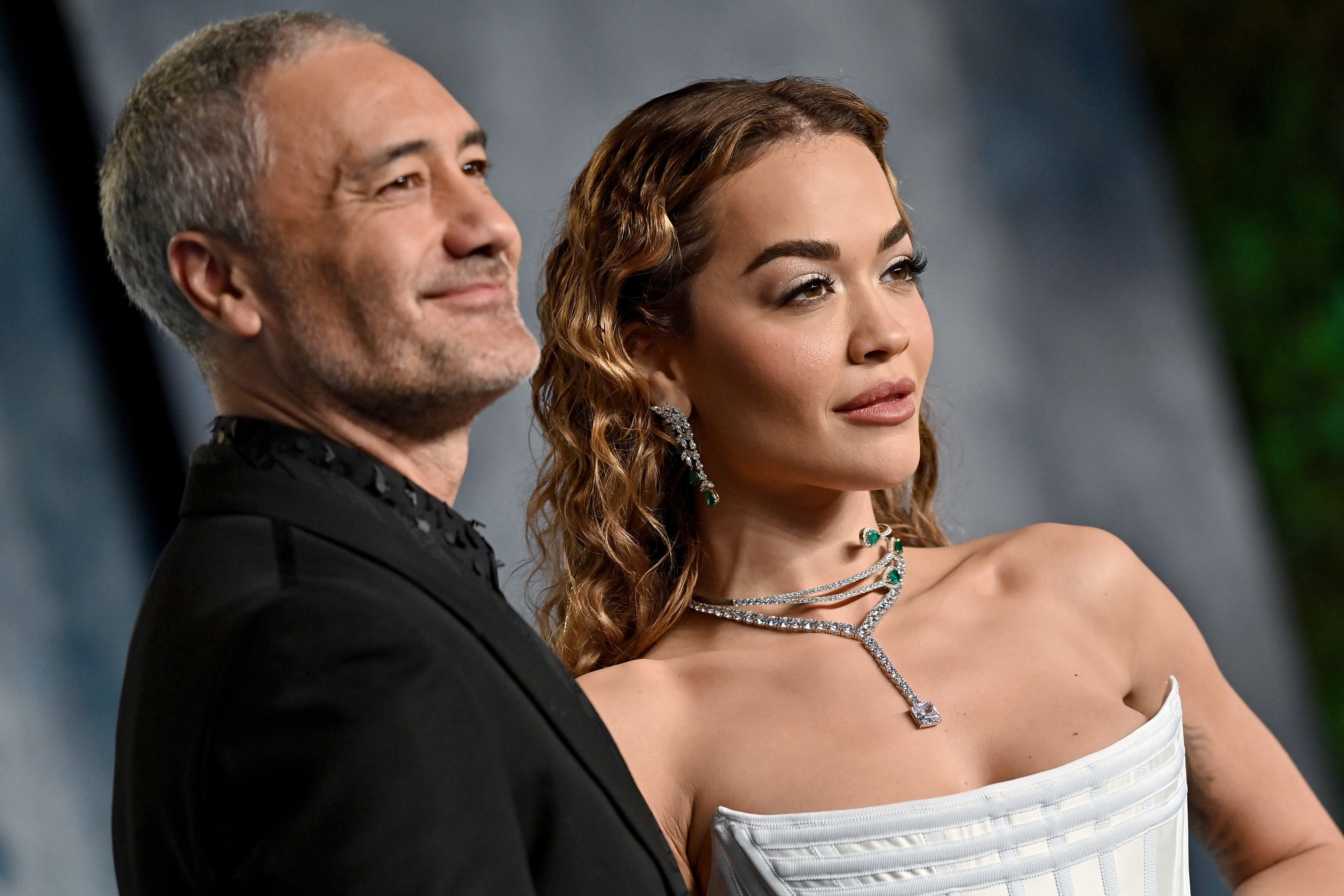 A close-up of Taika Waititi and Rita Ora posing together on the red carpet, he&#x27;s in a black suit and she&#x27;s in a white strapless gown