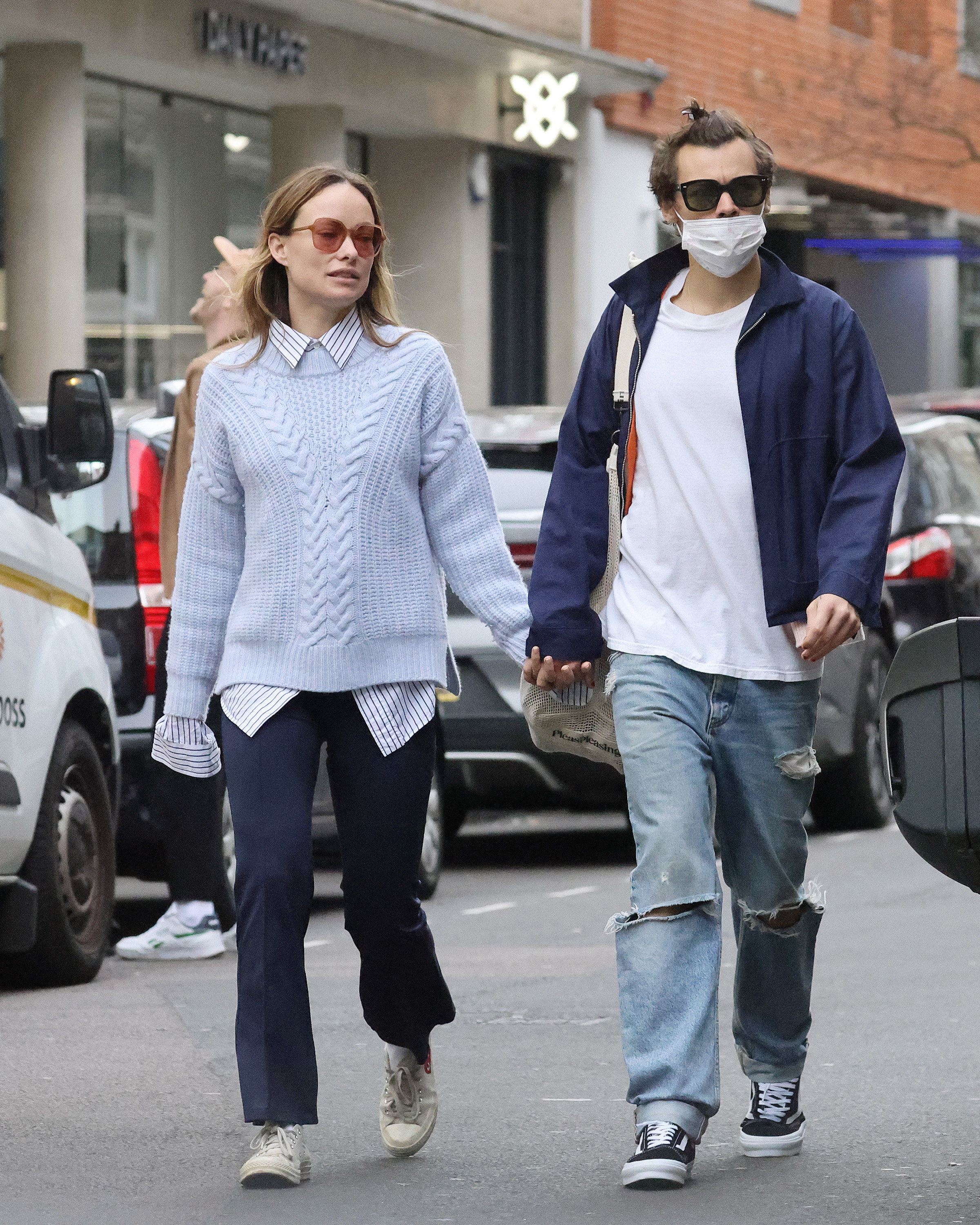 A paparazzi photo of Olivia Wilde and Harry Styles walking on the street hand in hand wearing casual clothes, jeans and sneakers, he&#x27;s wearing a mask