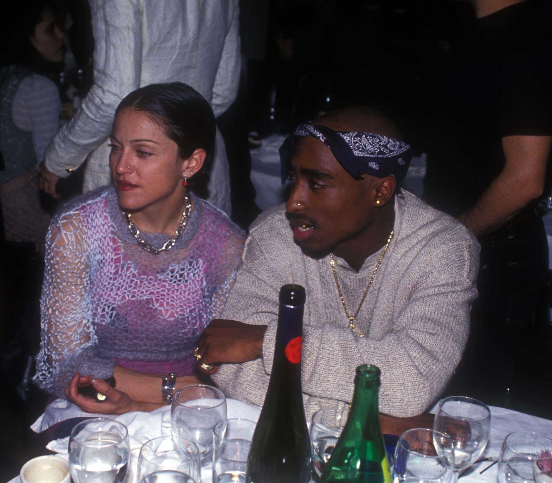 An old photo of Madonna and Tupac sitting at a table that&#x27;s covered in glasses and wine bottles, in conversation with someone to their right