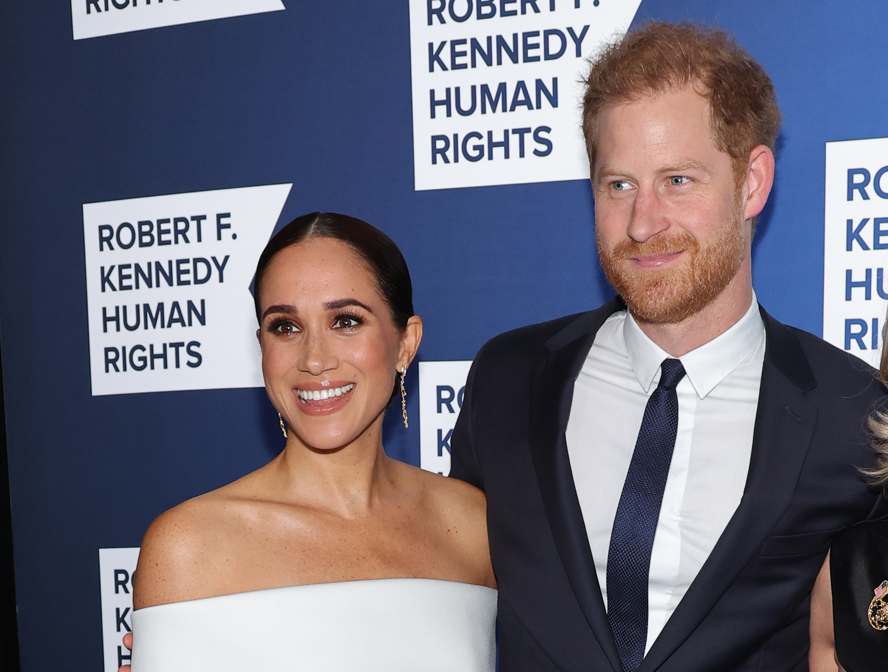 Meghan Markle and Prince Harry posing in front of a step-and-repeat, smiling and arm-in-arm