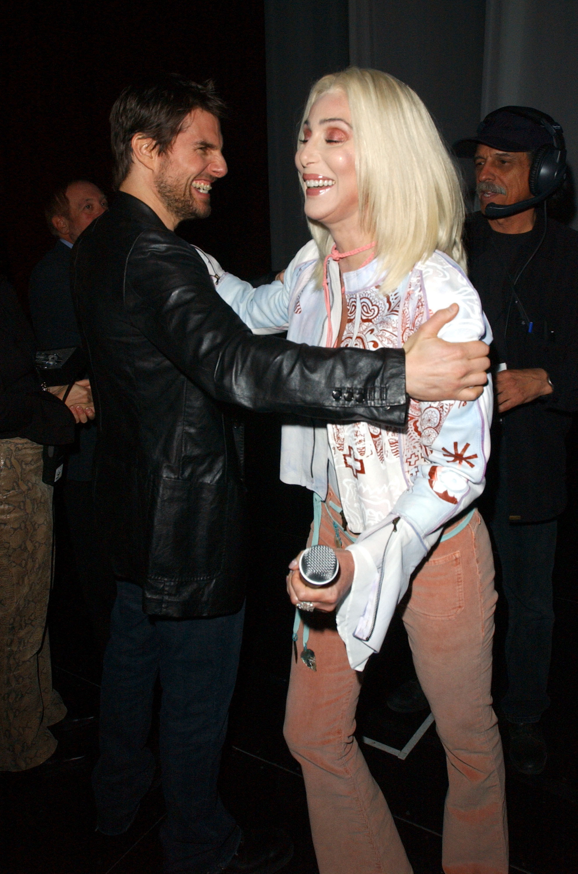 Tom Cruise and Cher in a half-embrace as she walks past him backstage, she&#x27;s holding a microphone and they&#x27;re both smiling