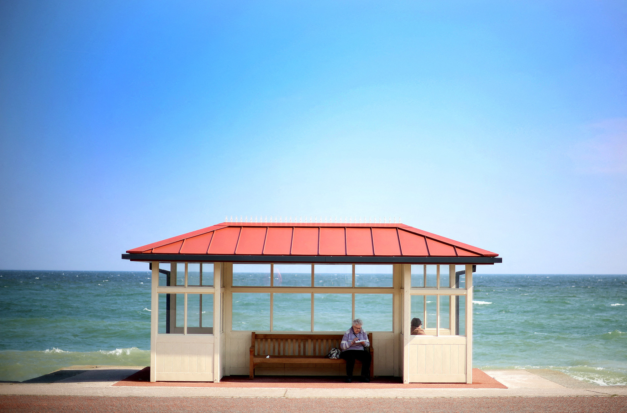 a woman sits on a bench inside a window-filled open shelter in front of the seashore