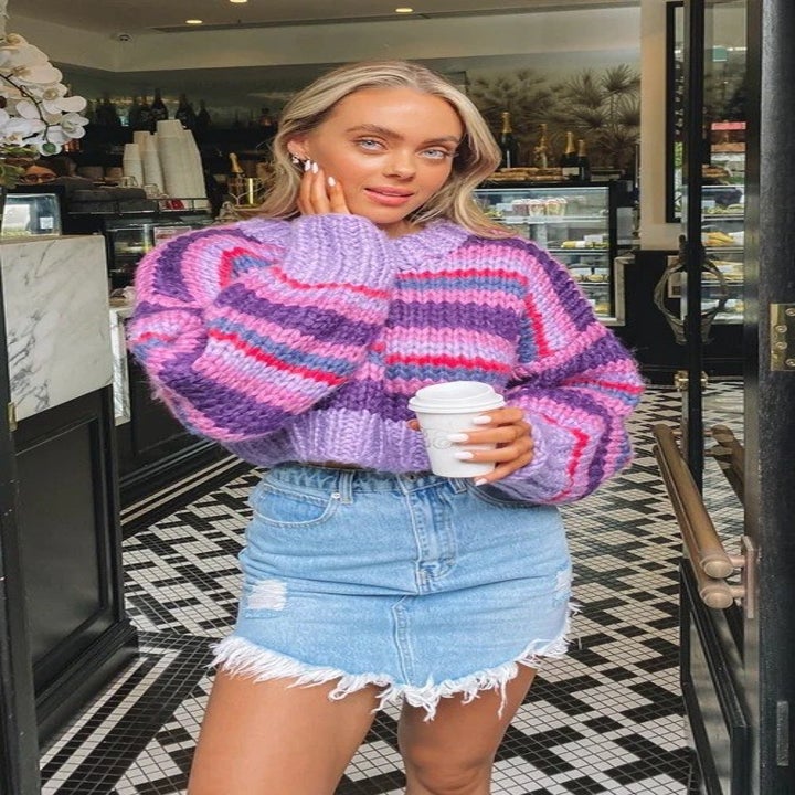 A model in a denim skirt and chunky striped sweater in shades of purple and pink