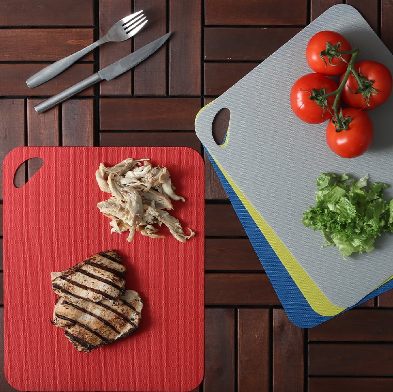 the red cutting board with chopped chicken on it, on a counter next to the blue, yellow, and grey boards. the grey board has tomatoes and chopped lettuce