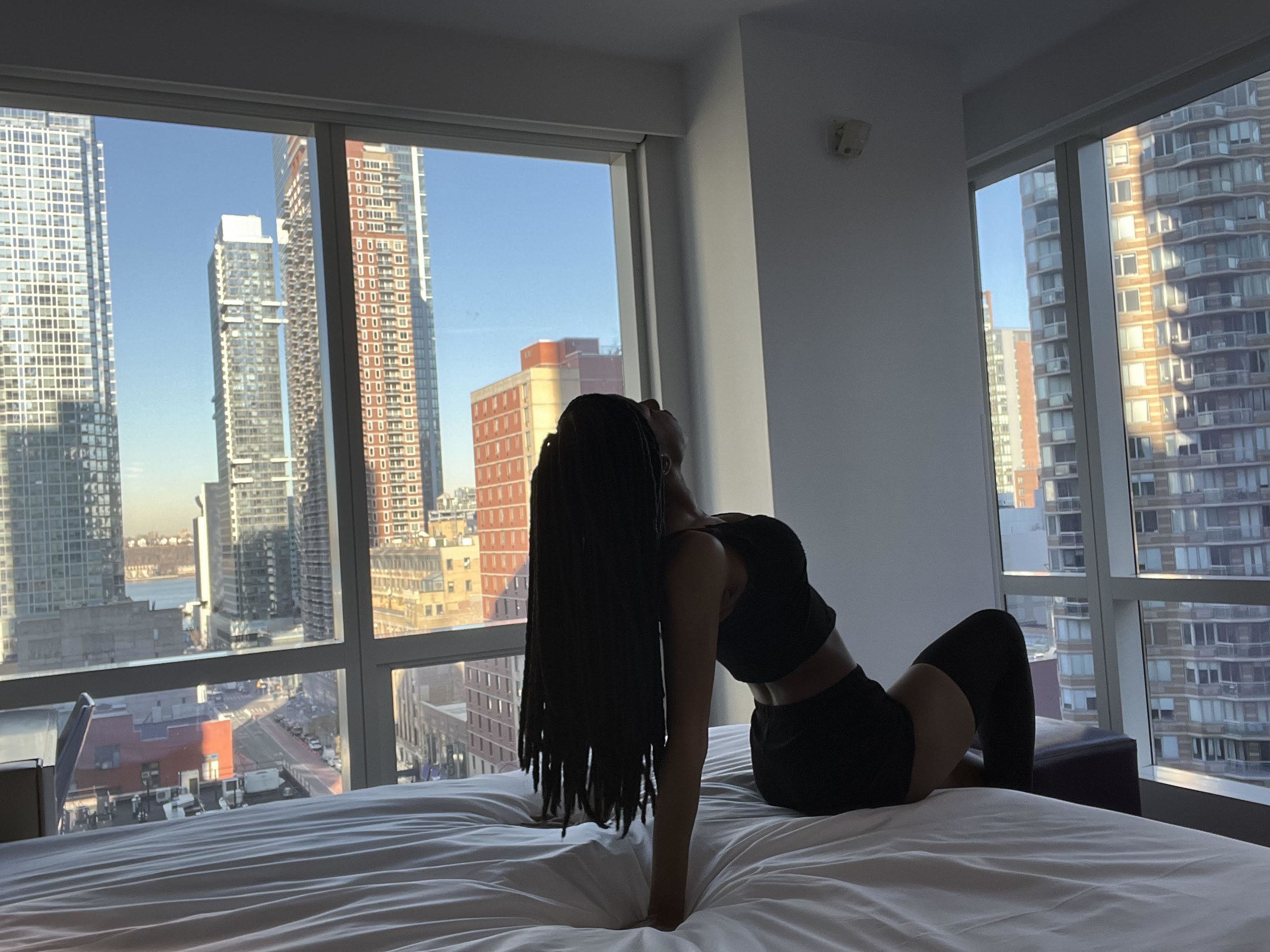 Woman wearing black lace two piece outfit while leaning on the corner of the bed inside of a hotel room
