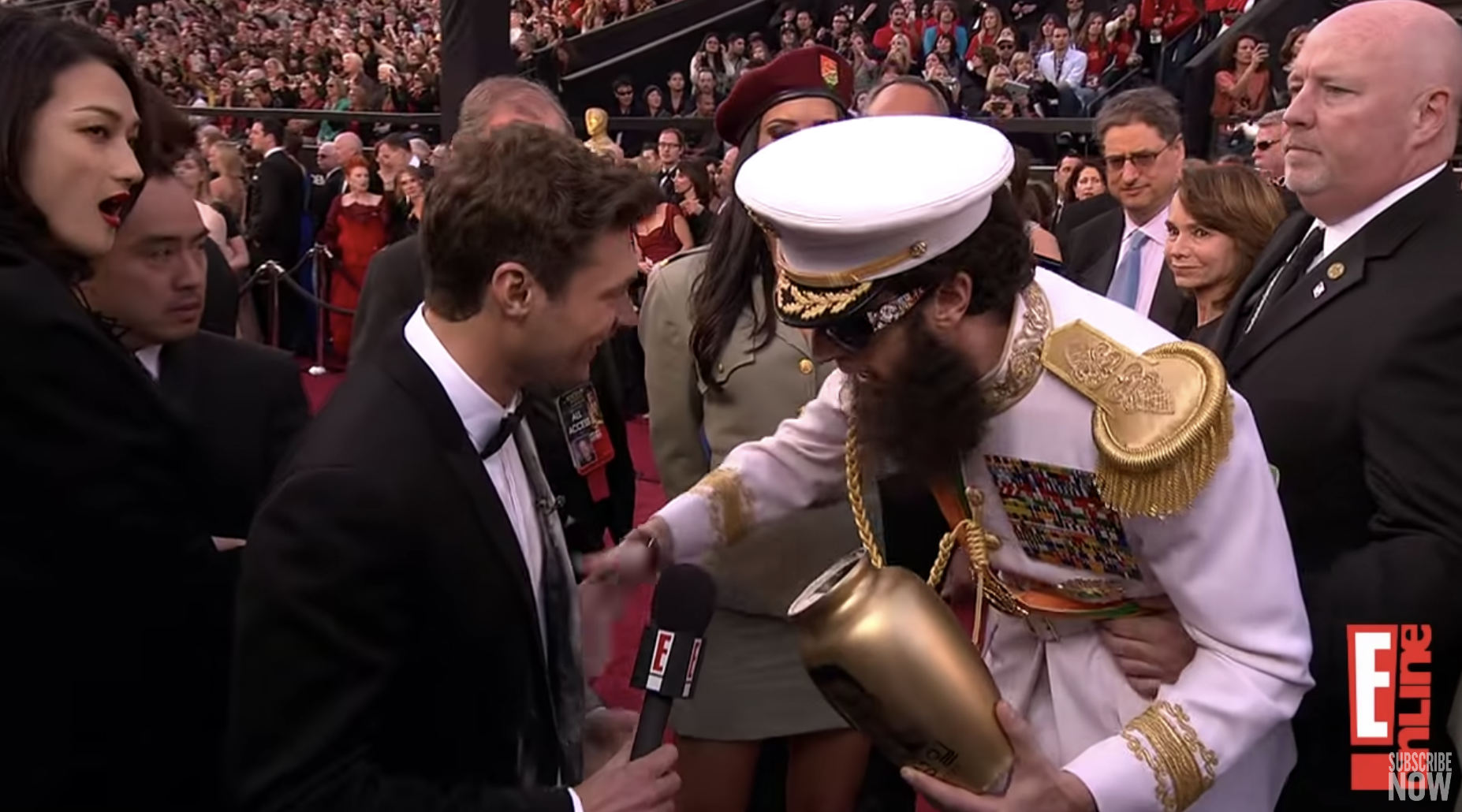 Sacha Baron Cohen, dressed in a white military uniform and holding an open urn, is wiping his hand on the front of Ryan Seacrest&#x27;s suit who is holding a microphone