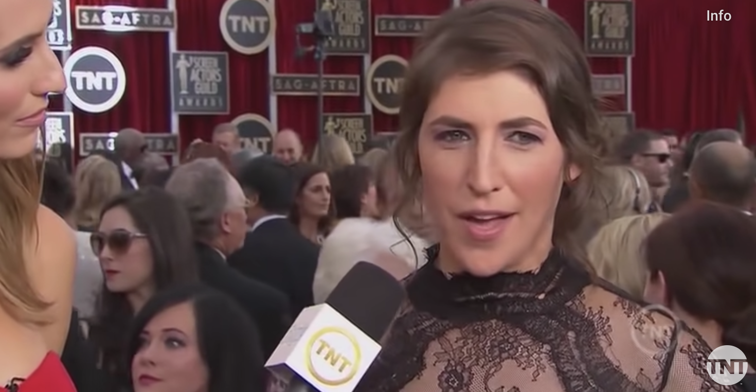 Mayim Balik wearing a lacy dress standing on the red carpet, with a lot of people behind her, being interviewed by a woman with a microphone