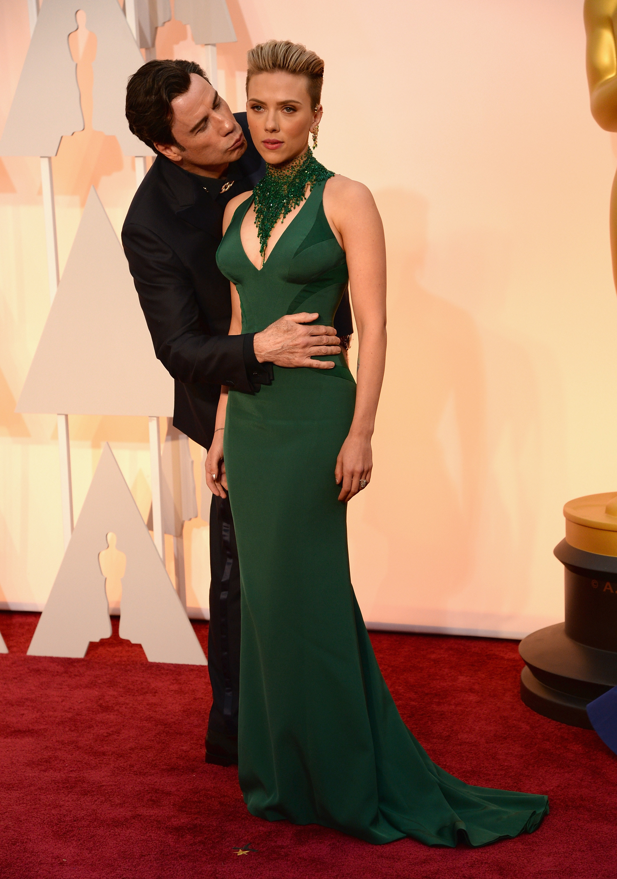 Scarlett Johansson stands on the Oscars red carpet in a green gown, John Travolta stands behind her with his hand on her stomach, he&#x27;s leaning in to kiss her cheek