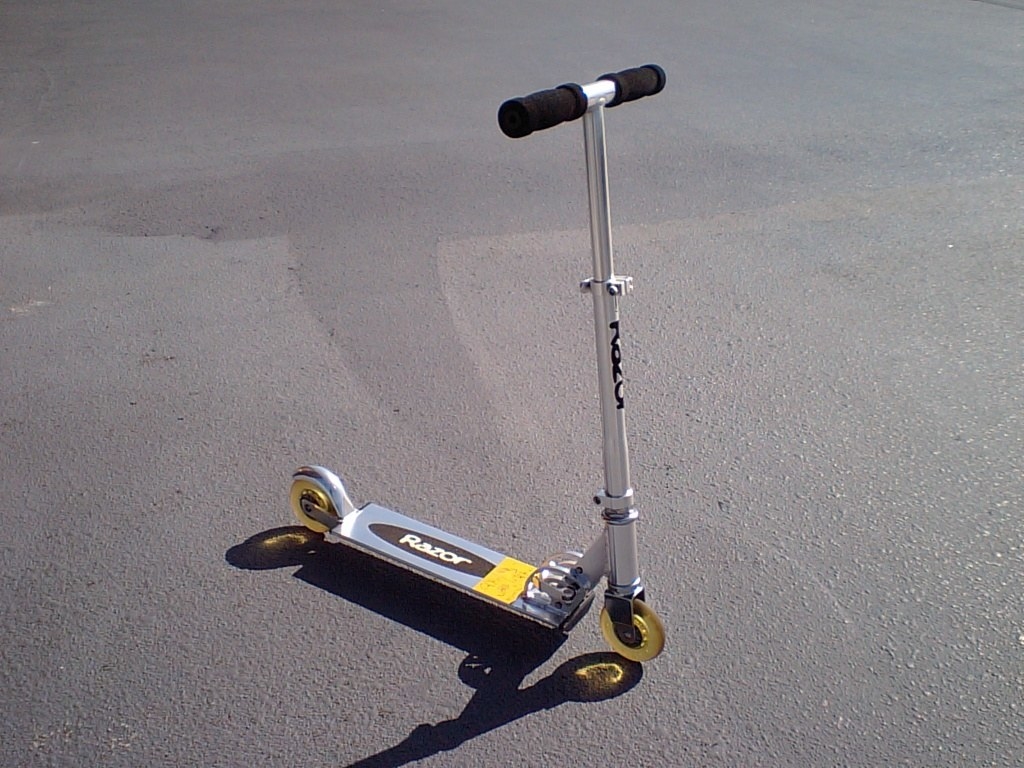 A two-wheeled handlebar scooter standing up straight on pavement. Not sure how it&#x27;s standing up on its own