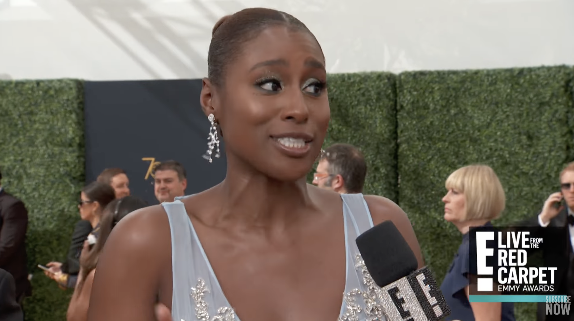Issa Rae stands on a red carpet, wearing a blue sleeveless gown and long earrings, there&#x27;s a microphone in front of her