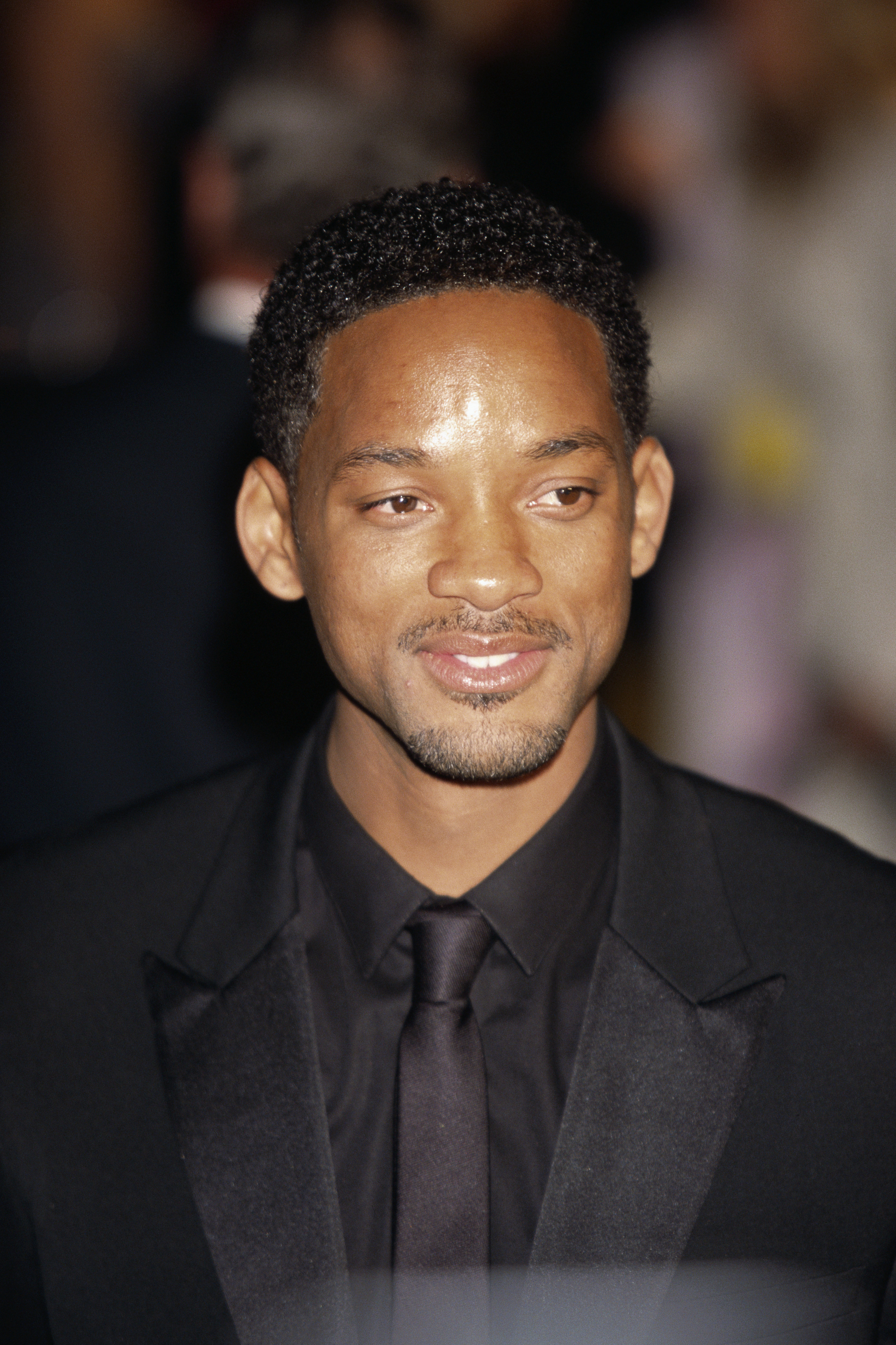 Will Smith smiling on a red carpet in a black suit with black tie