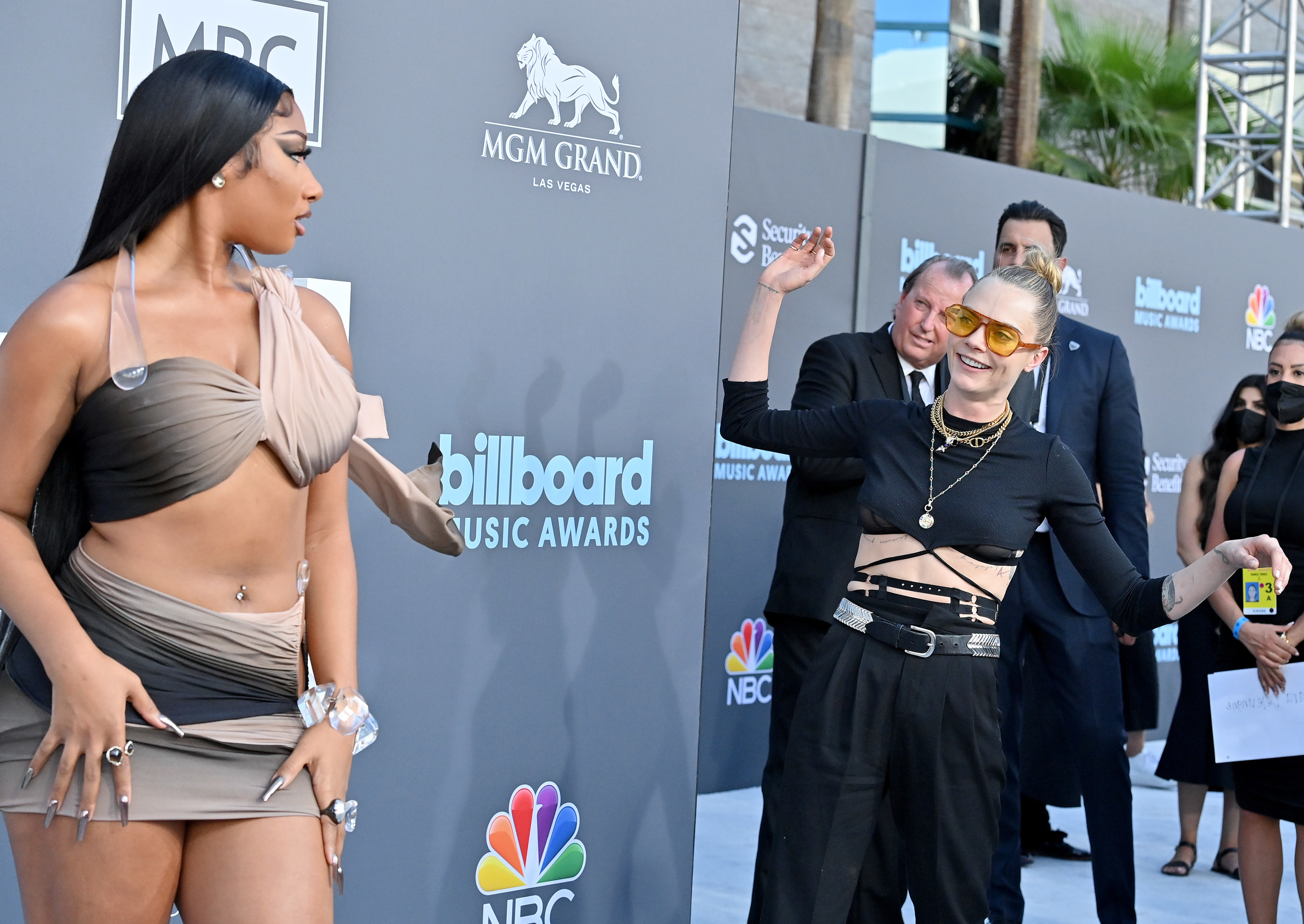 Cara Delevingne is standing on the Billboard awards blue carpet, smiling with her arms up while looking at Megan Thee Stallion