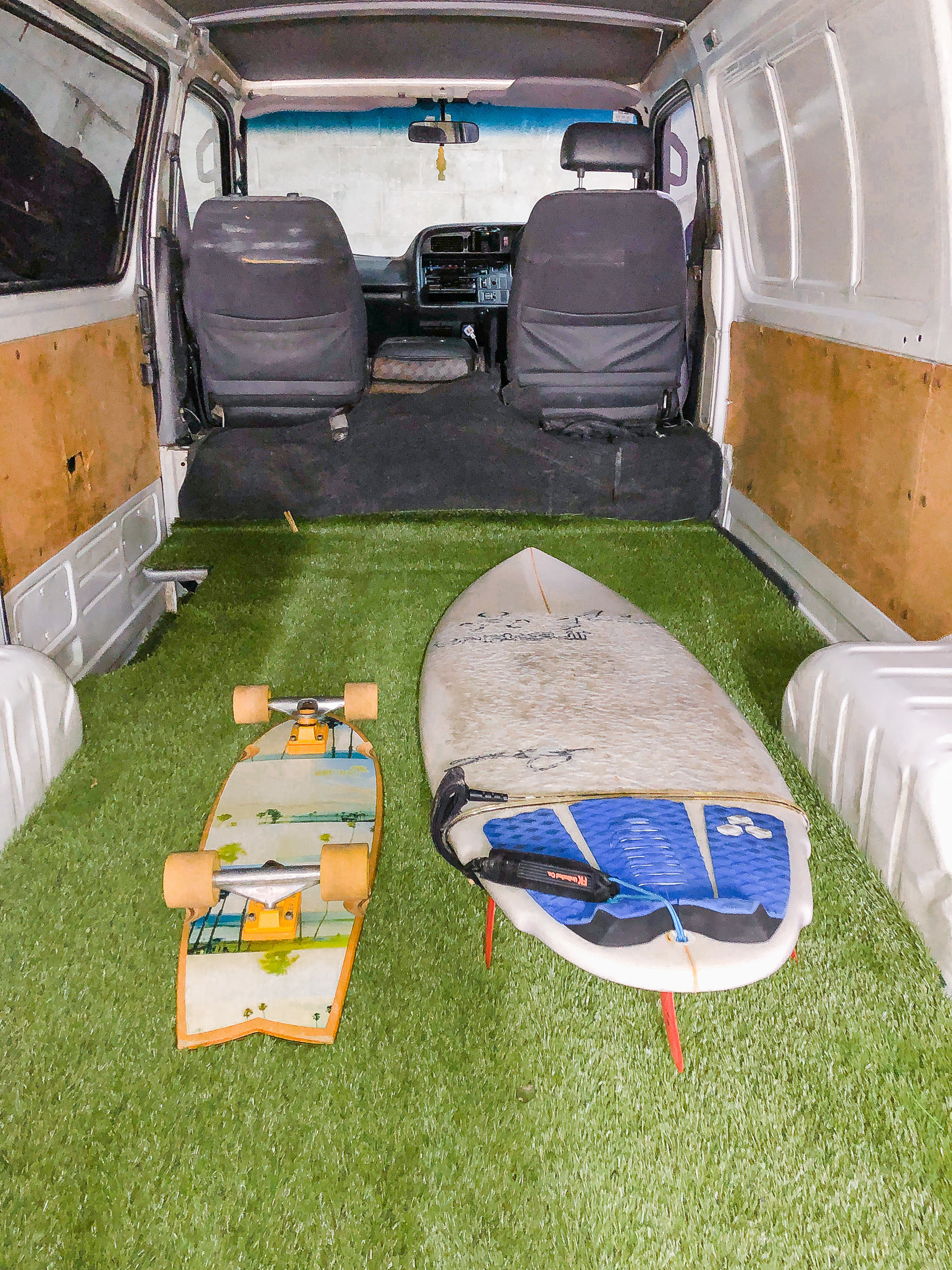 Photo of empty van with grass in the back, a skateboard, and a surfboard