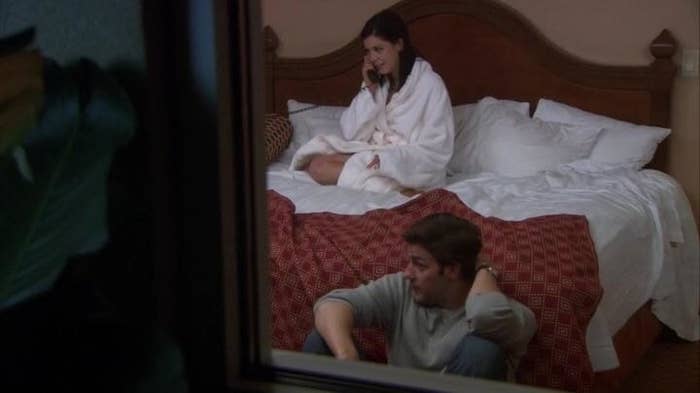 Cathy sitting on the bed in a bathrobe on the phone while jim sits on the floor looking uncomfortable