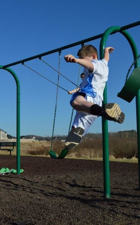 A young boy in mid-air jumping off a swingset. Another person swings in the back