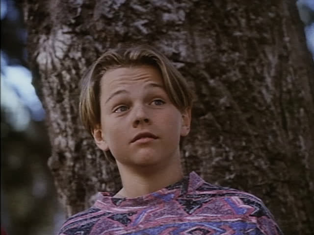 A young and round-faced Leo DiCaprio stands in front of a tree looking surprised