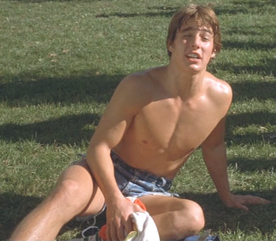 A young Tom Cruise sits on the grass, shirtless in denim cutoff shorts